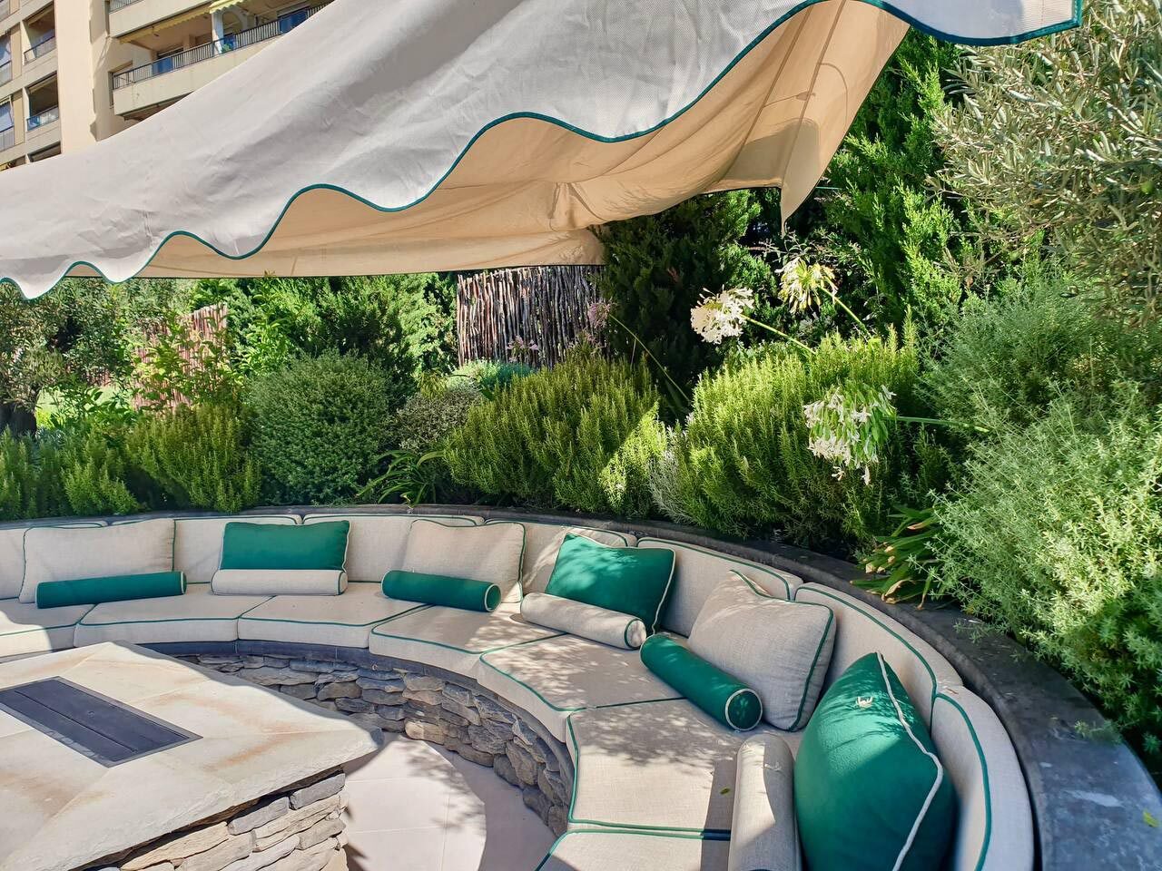 Salon extérieur circulaire en pierre avec coussins verts sous voile d'ombrage beige, jardin méditerranéen
